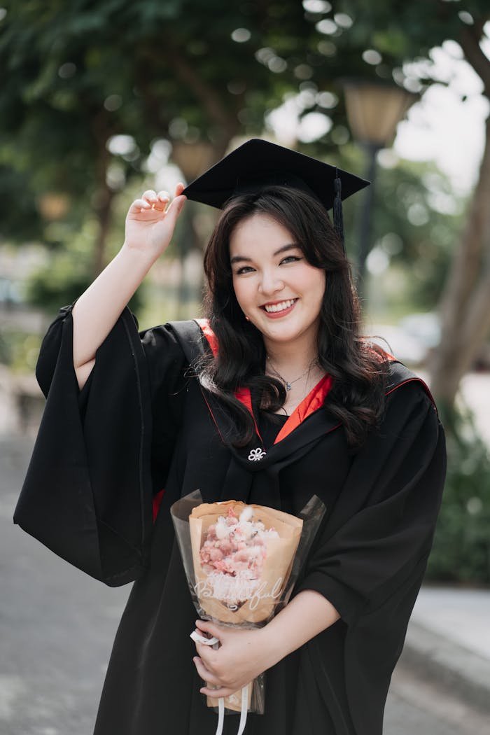 Smiling graduate in cap and gown holding flowers outdoors, celebrating achievement.