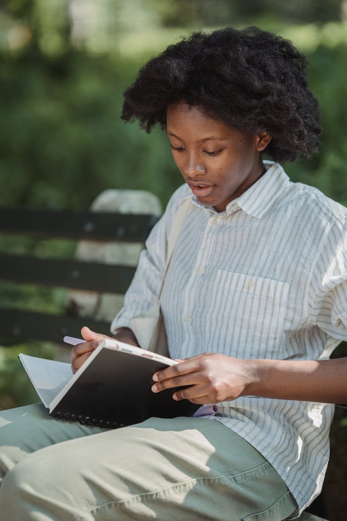 A young woman seated on a park bench writing in a notebook, surrounded by nature.