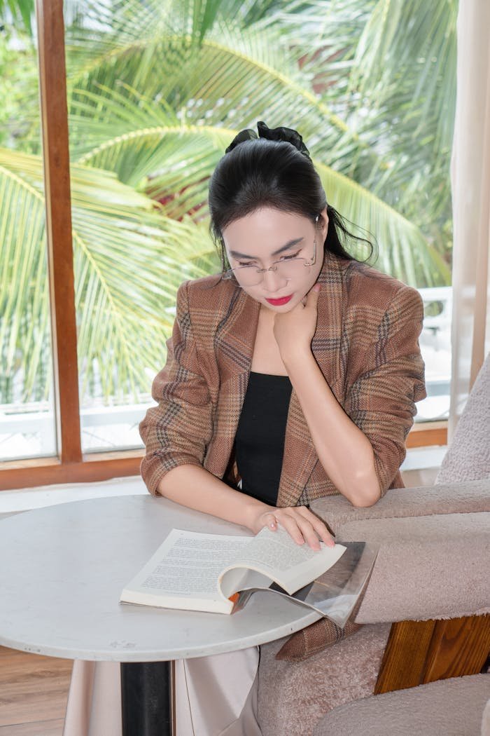 A woman in glasses reads a book indoors with tropical palm trees outside the window.