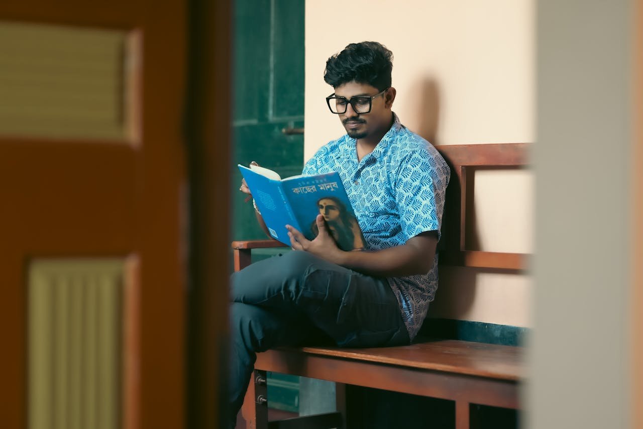 A young man sitting on a bench reading a book indoors. Calm and focused study environment.